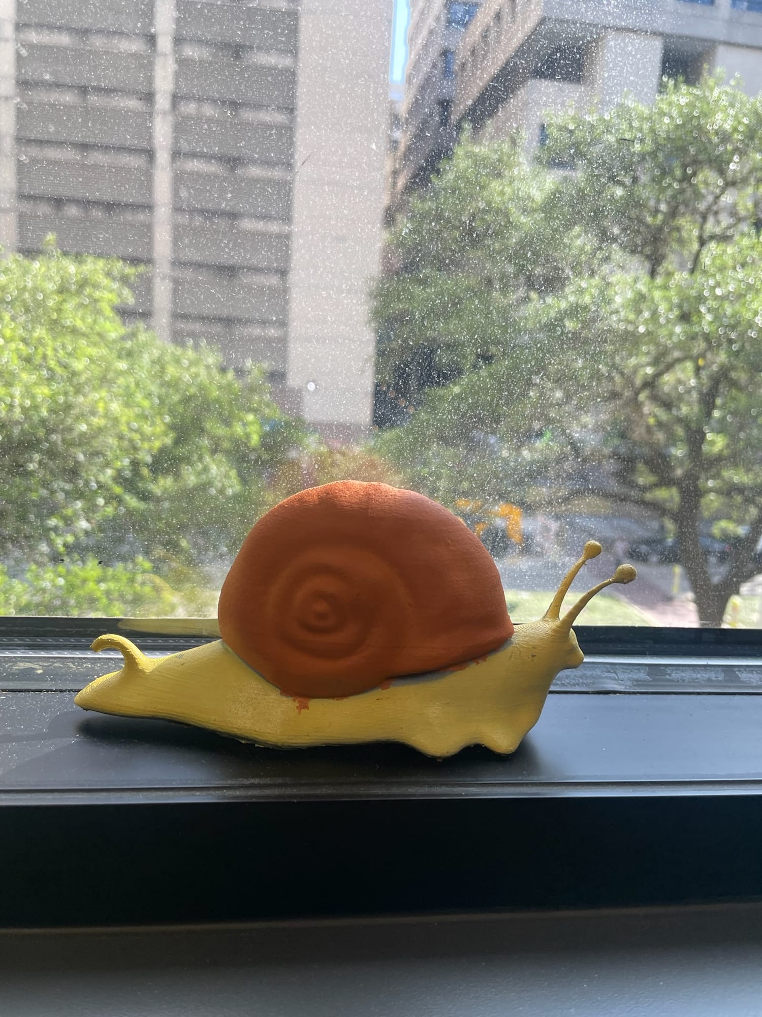 A 3D-printed snail with a painted red shell sits on a windowsill overlooking trees