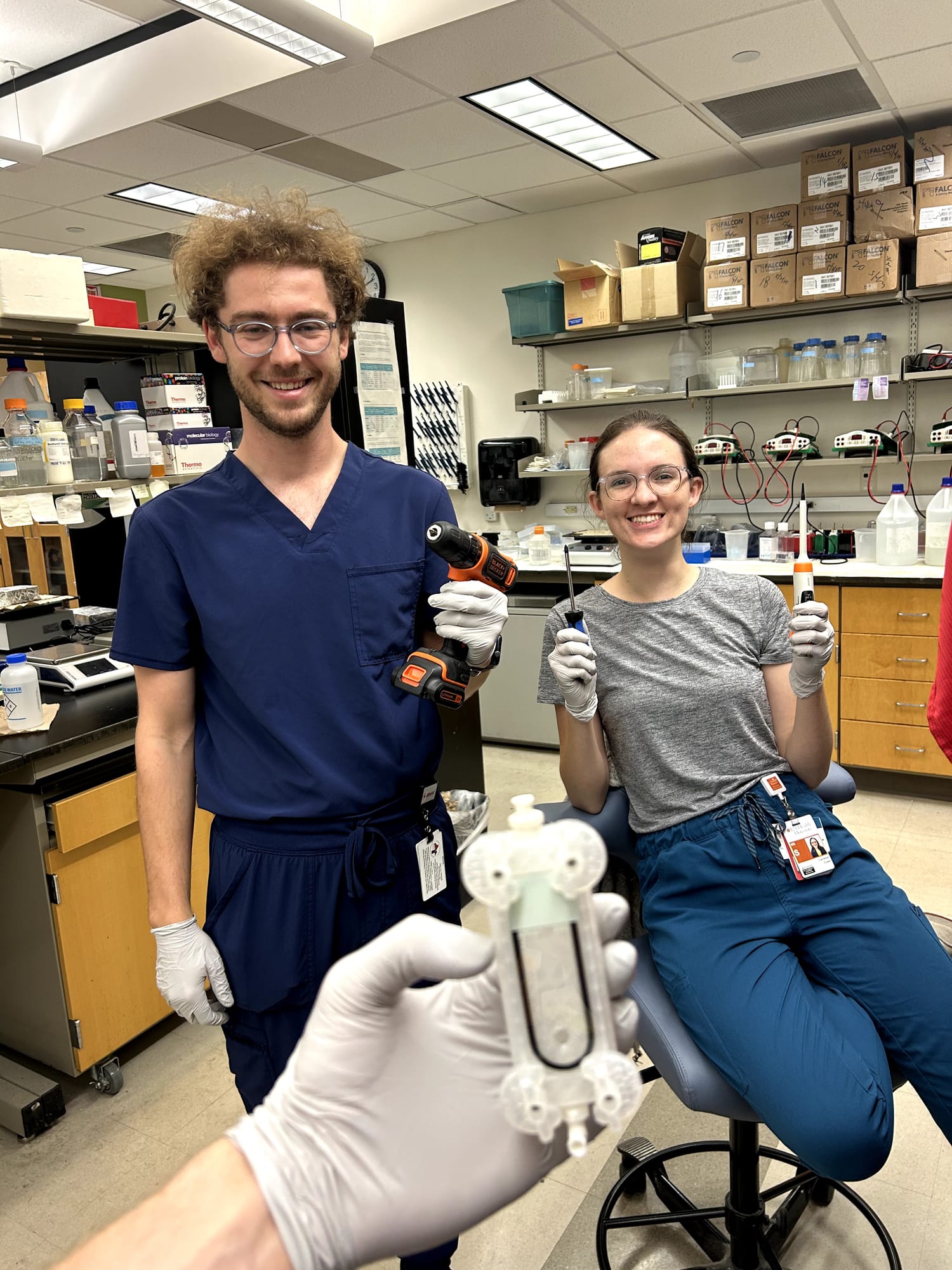 Two researchers smile in the lab, one holding the prototype in a gloved hand