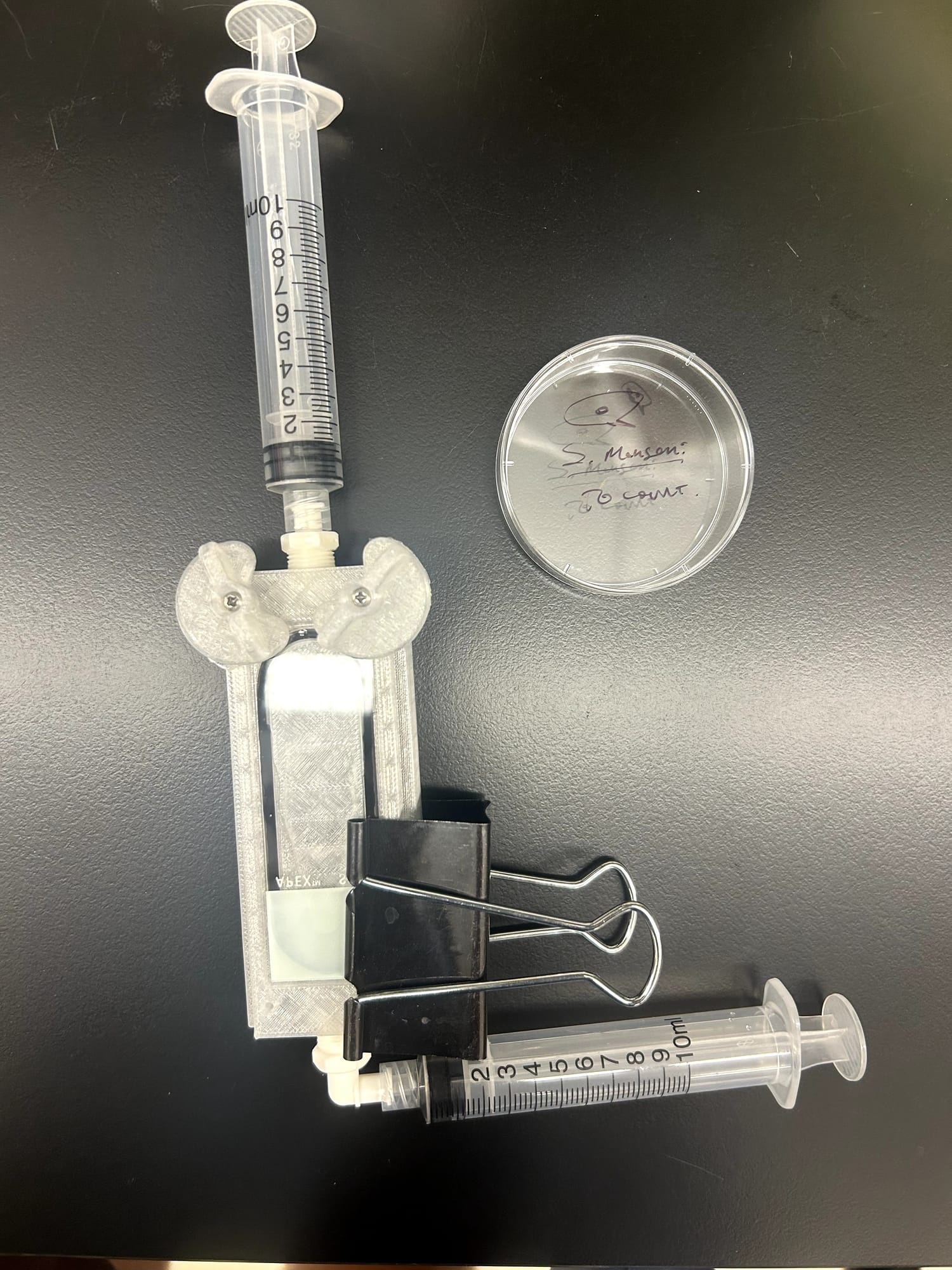 A 3D-printed prototype with syringes and a binder clip beside a small petri dish