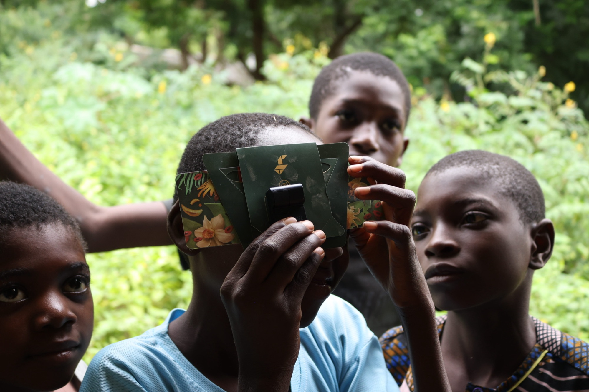 Four children gather outdoors as one holds a Foldscope paper microscope up to one eye like a camera.