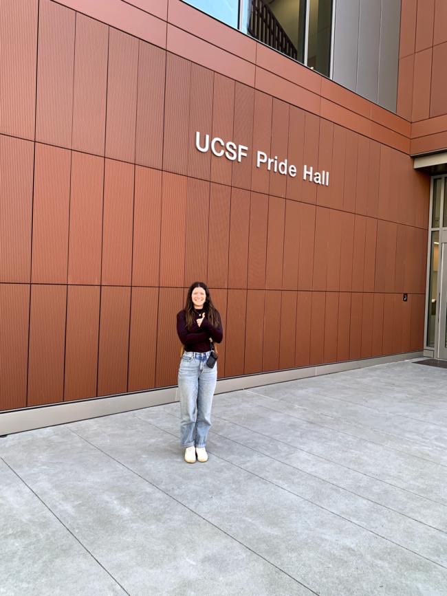 Maya stands smiling in front of a wall reading 'UCSF Pride Hall'