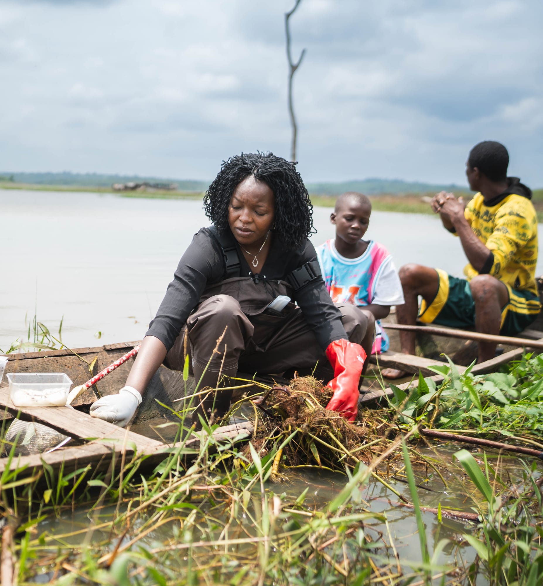 Dr. Olubukola Adelakun kneels in a boat pulling snails from a mass of aquatic plants, with community members seated behind her