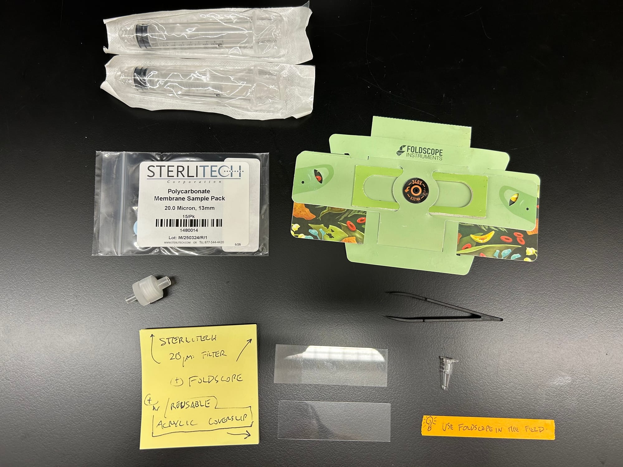 Flat lay on a black bench: syringes, a Sterlitech filter pack, a green Foldscope, glass and acrylic slides, and a sticky note listing the parts