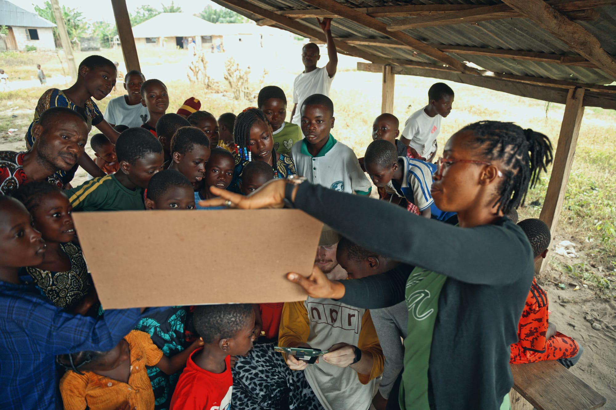 An educator holds up a card to a crowd of schoolchildren gathered under an open-sided shelter