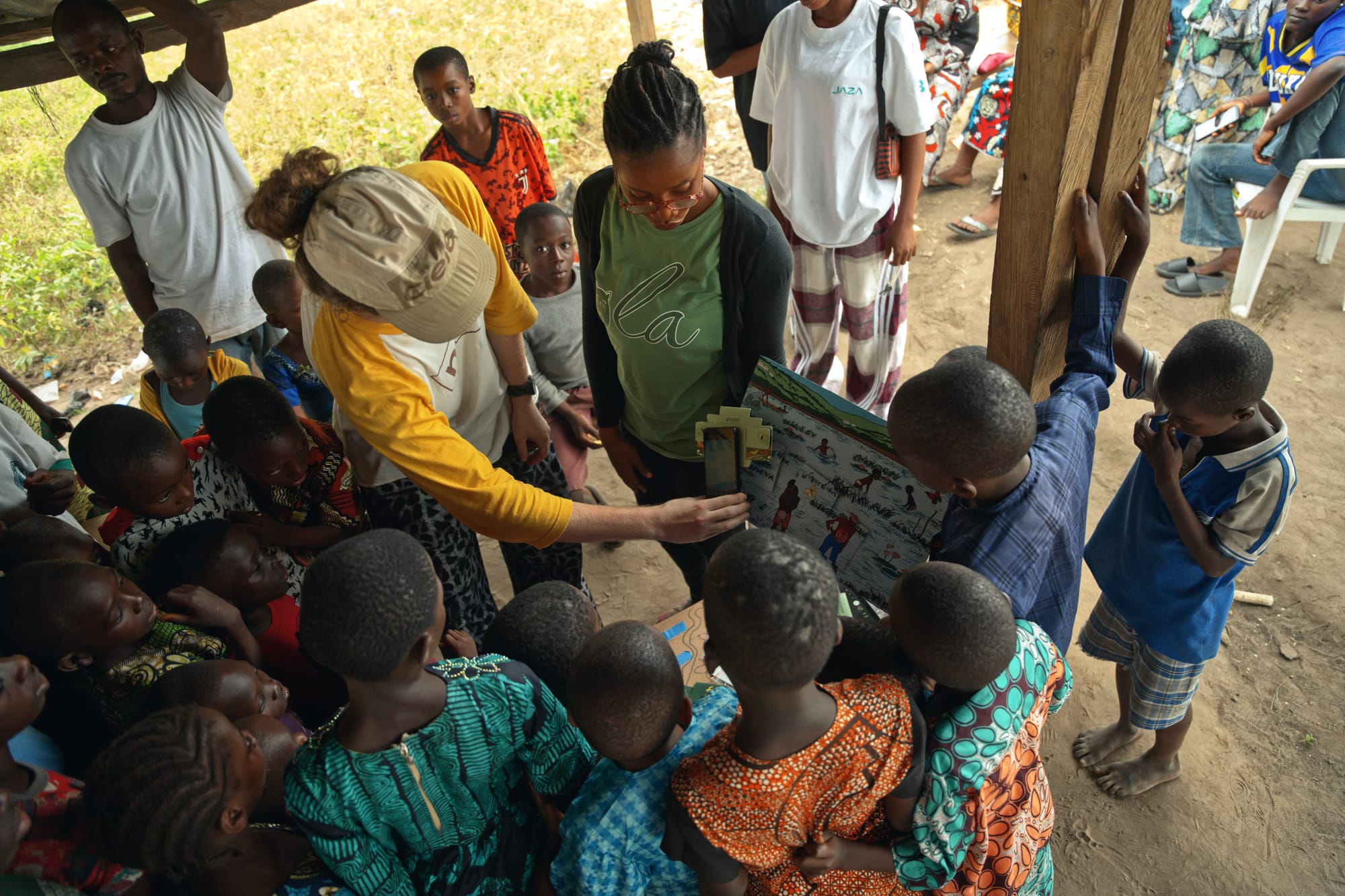 Children and adults crowd around an educator in a green shirt and a team member in a straw hat, who hold up puzzle pieces under a shelter