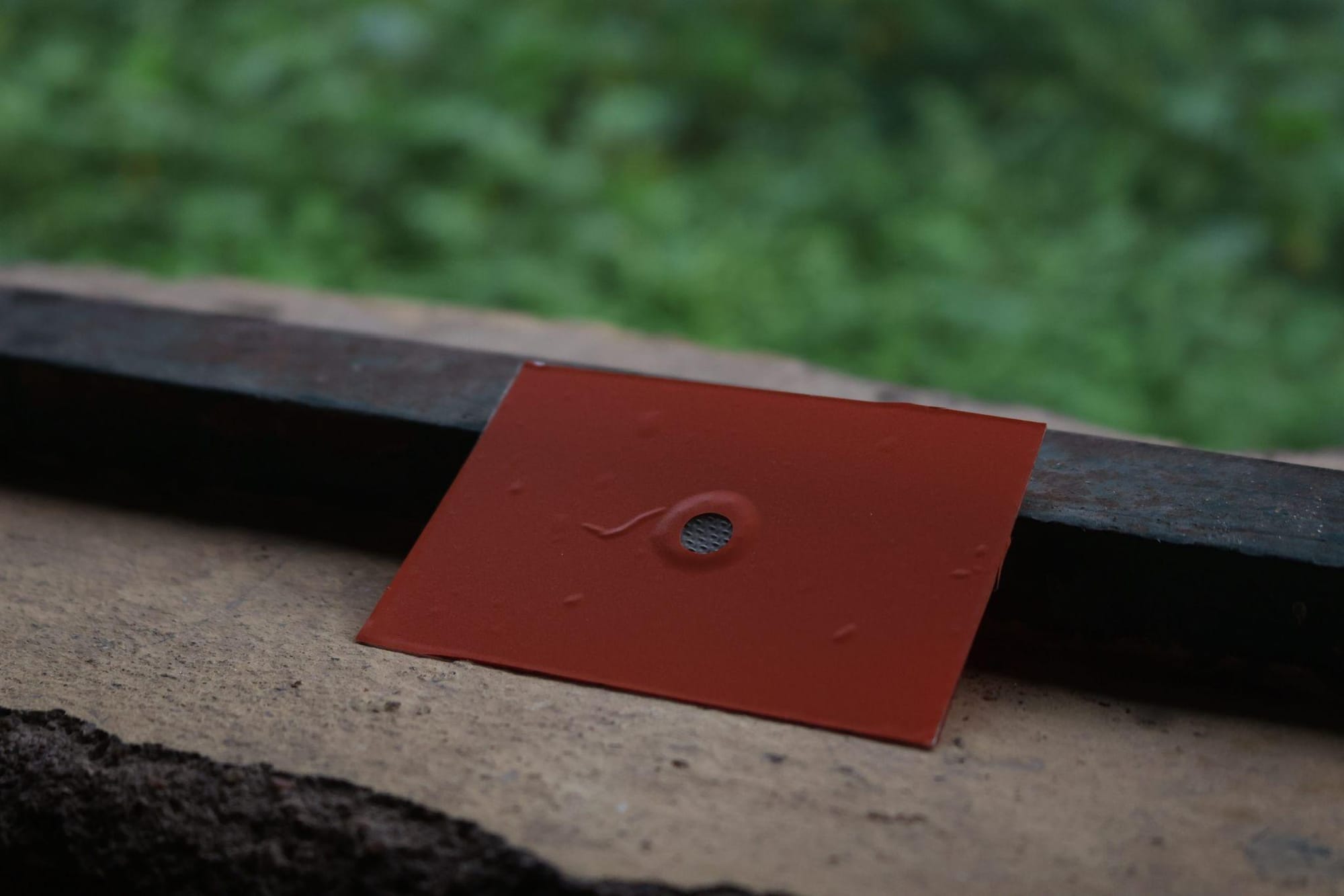 An orange SchistoFilter card with a small stainless-steel mesh window, leaning on a wooden ledge