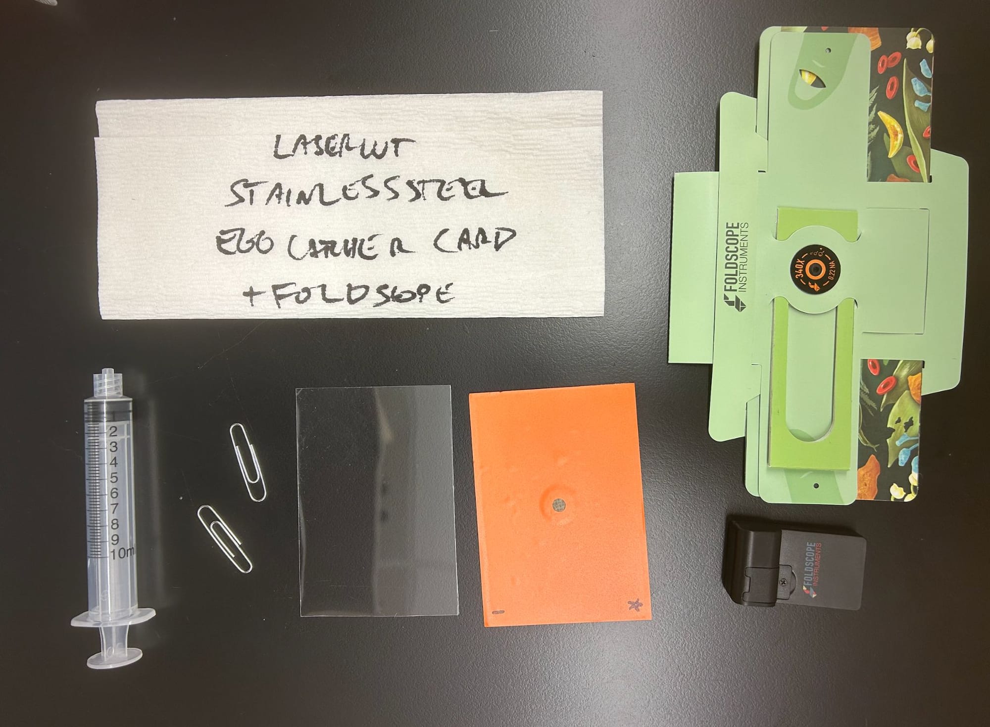 Flat lay of a syringe, paperclips, an acrylic cover, an orange laser-cut filter card, a Foldscope, and a napkin labeled lasercut stainless steel egg catcher card plus Foldscope