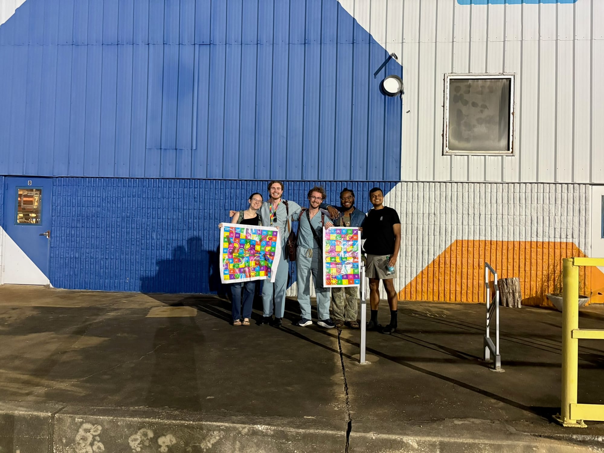 Five team members stand outside a blue-and-white makerspace building holding colorful posters