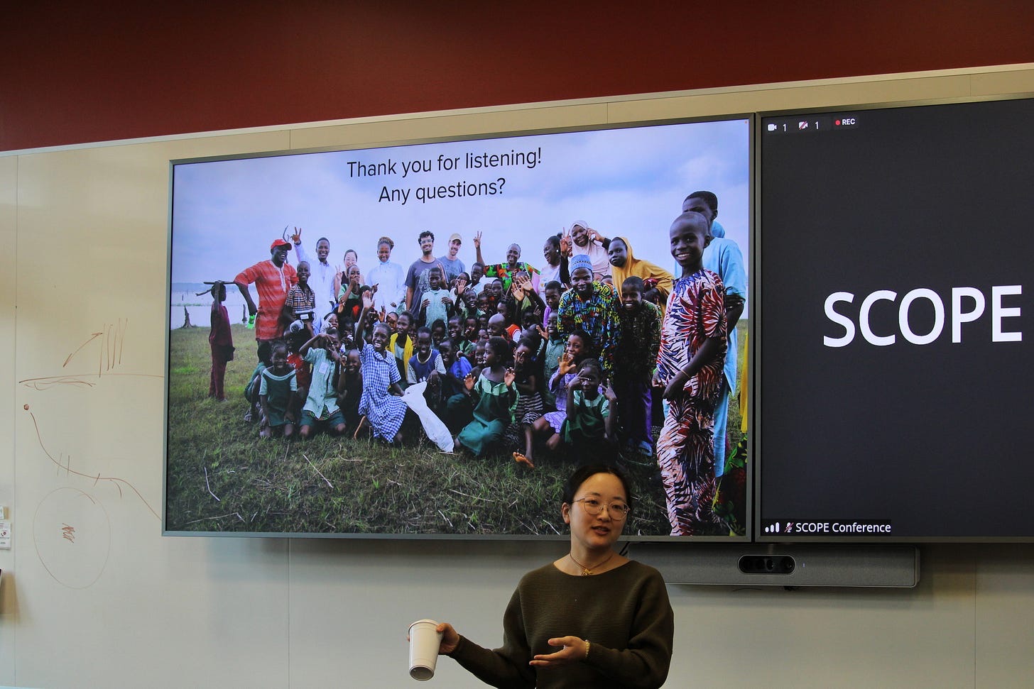 A presenter standing beside a projector screen addressing a room of seated students and faculty