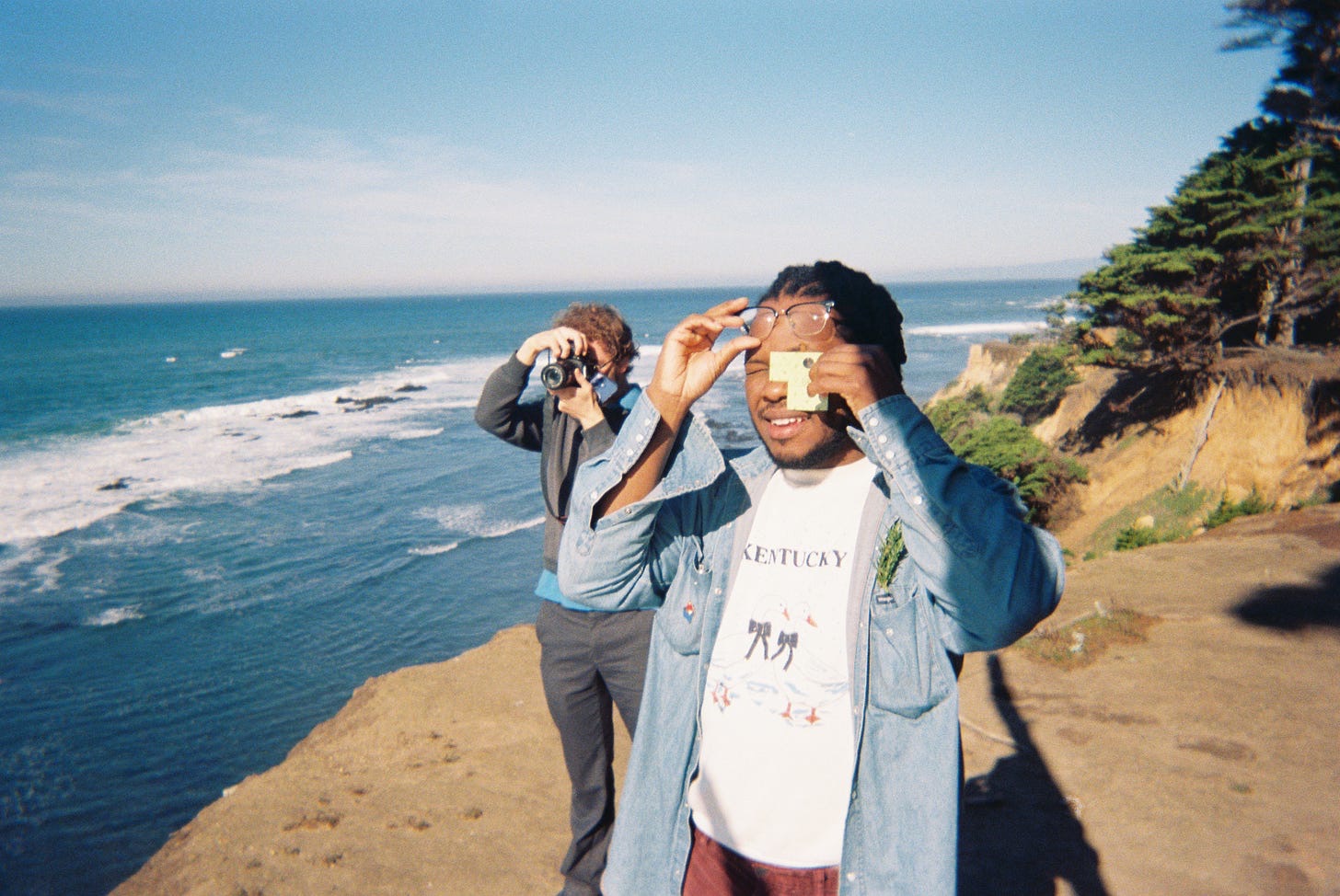 Two students crouched on rocks at the ocean’s edge, one looking through a camera and the other through a small paper microscope