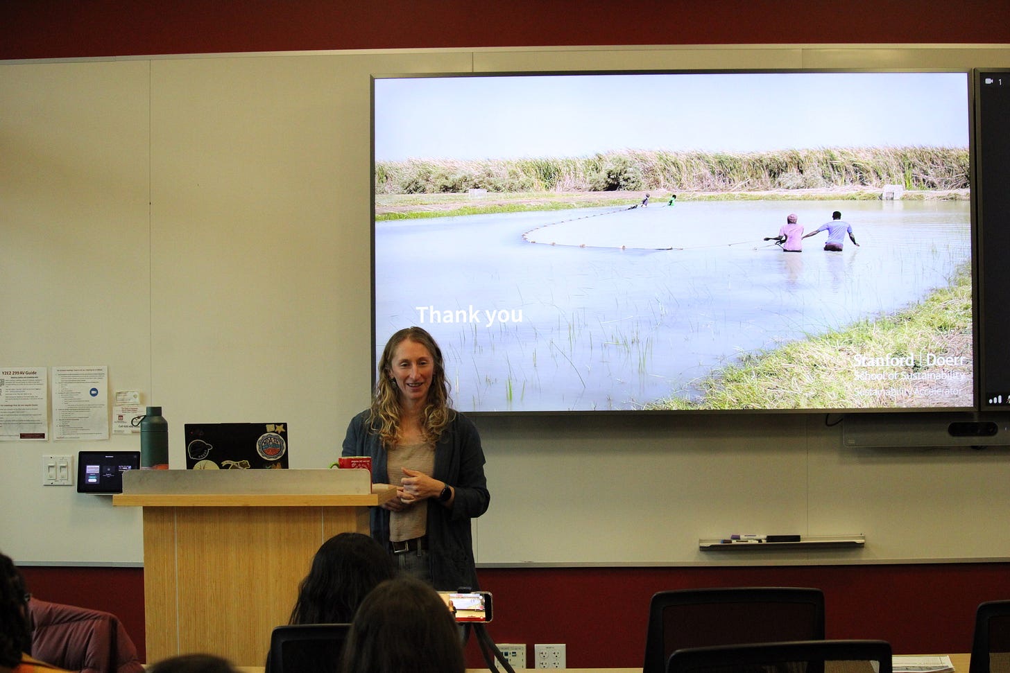 A speaker presenting in front of a screen showing people seining a pond, to a seated audience