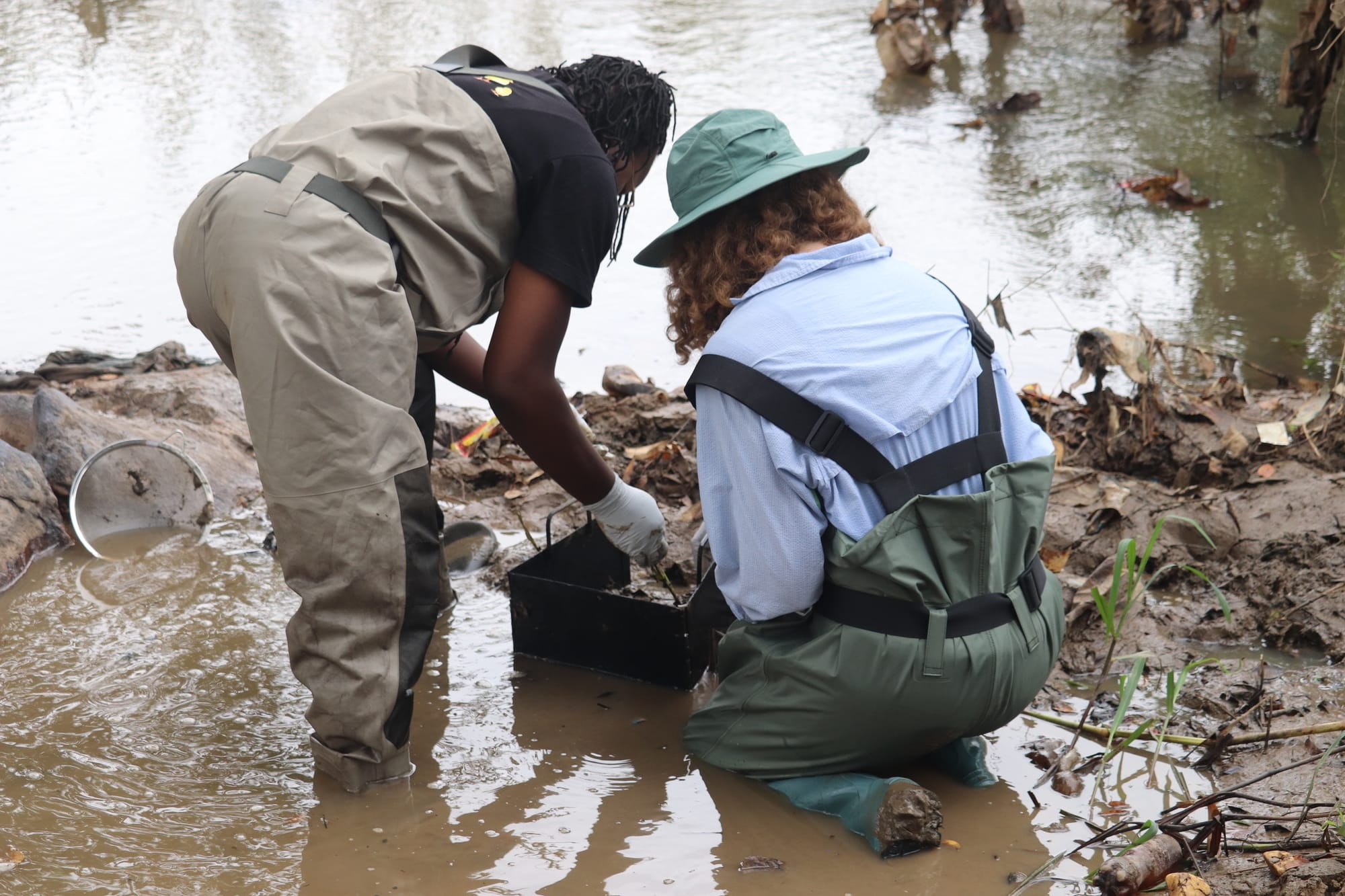 Two researchers in waders crouch in shallow water, one lowering a black welded metal quadrat onto the riverbed