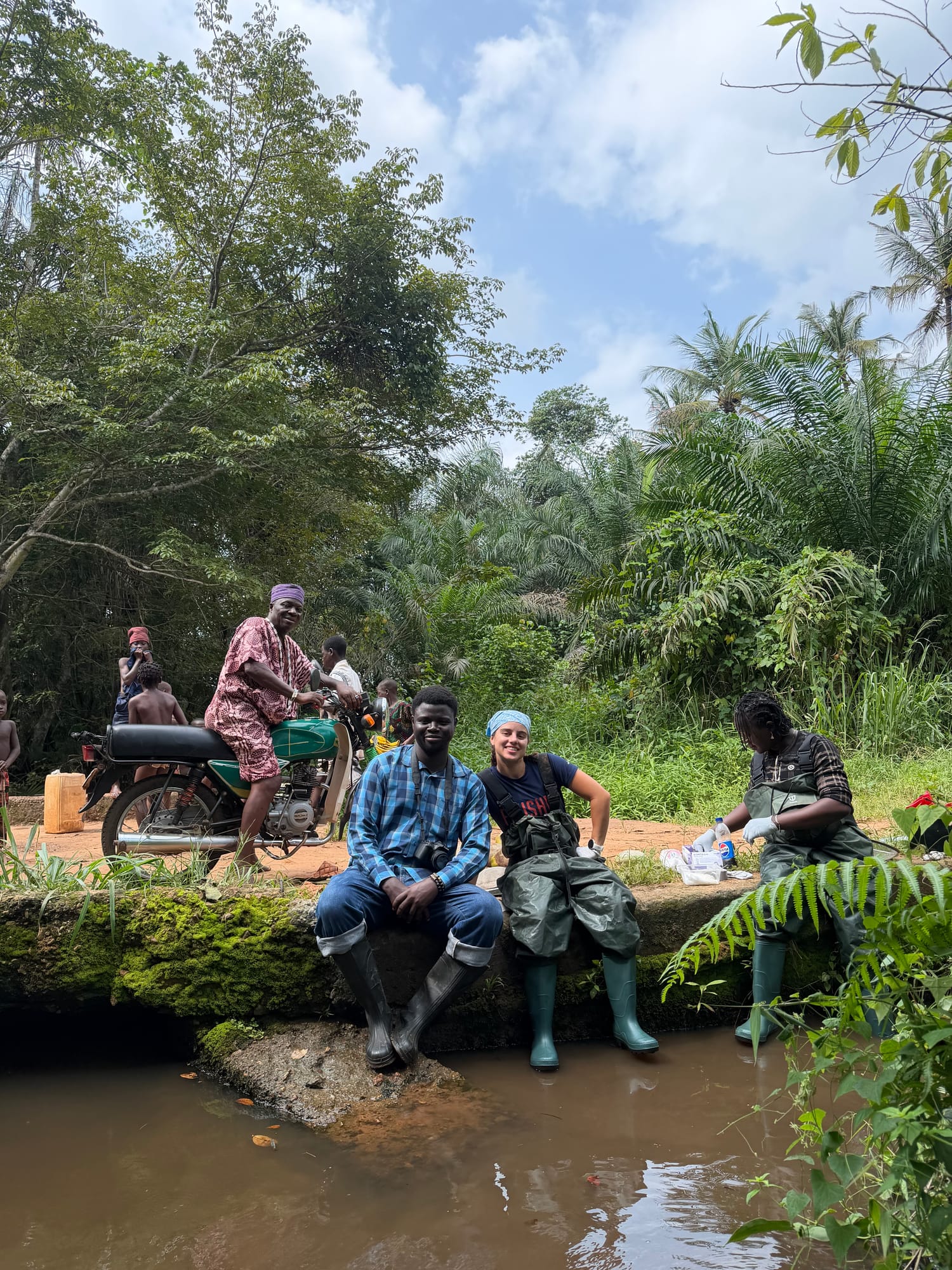 Community members sit on a mossy log at the river’s edge beside a motorbike, watching the survey team