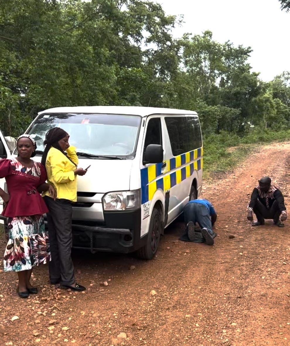 The team waits beside the van on a dirt road while one person kneels to look underneath it